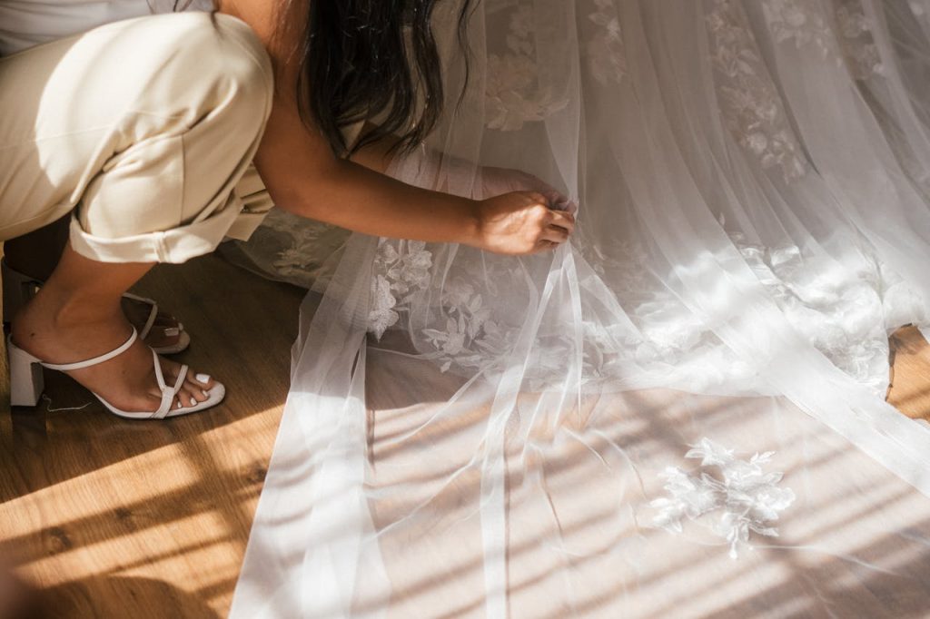 A beautiful brides veil and dress detail being adjusted indoors, capturing the essence of a wedding day.