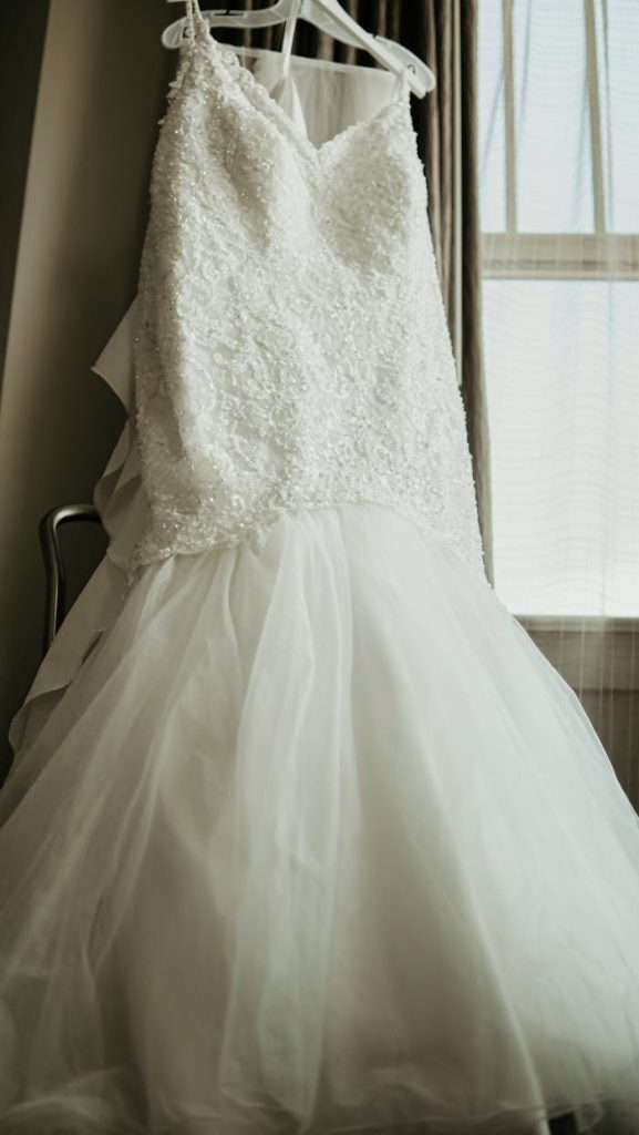 Stunning white wedding gown with lace and tulle details hanging indoors by a window.